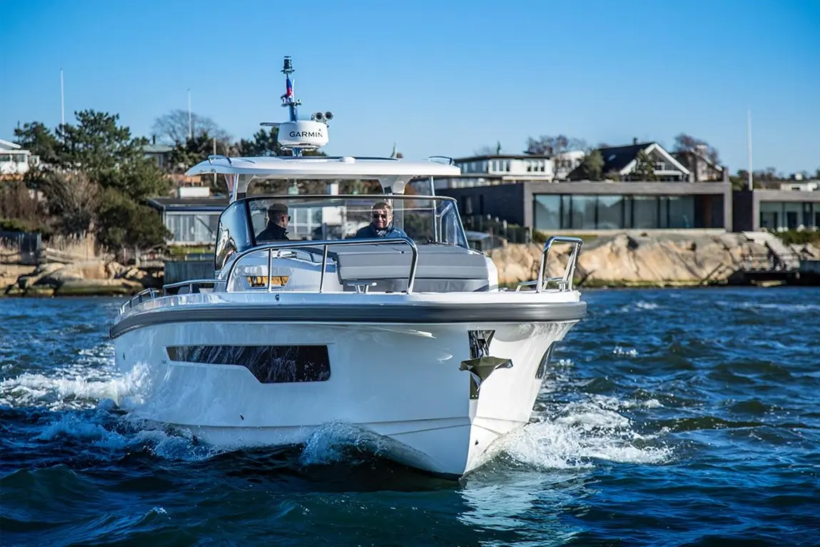 A white motorboat with two people onboard navigates through blue water, showcasing a waterfront with modern houses.